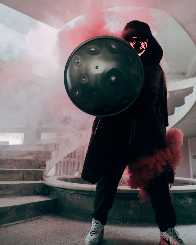 A mysterious performer plays a handpan in an urban setting, surrounded by vibrant smoke and neon lights.