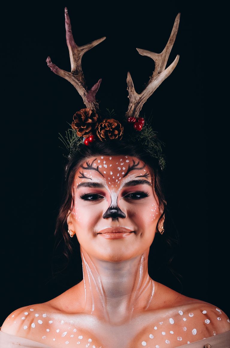 Artistic portrait of a woman with deer makeup and antler headband against dark background.